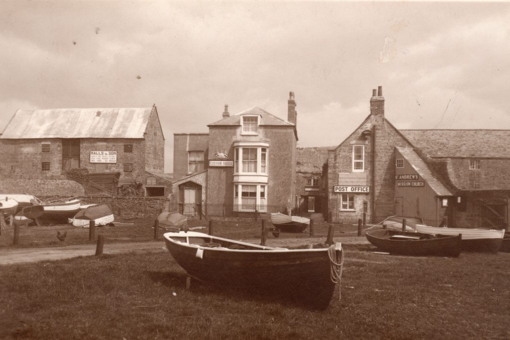 Old image of Bridport harbour showing boats laid up on the grass and buildings in the background.